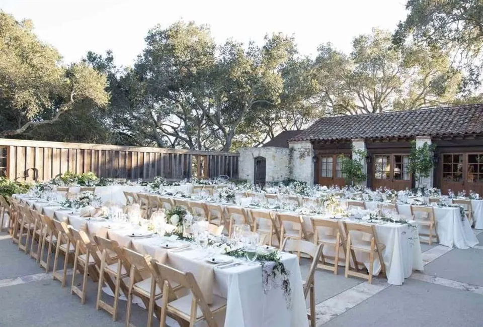 Long white tables set outdoors in a courtyard with wooden chairs